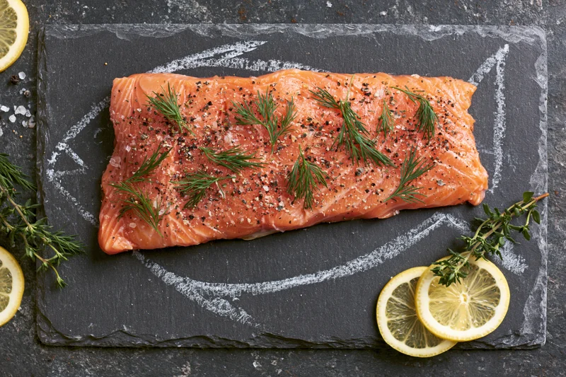 Raw salmon fillet seasoned with dill, pepper, and lemon slices on a slate plate