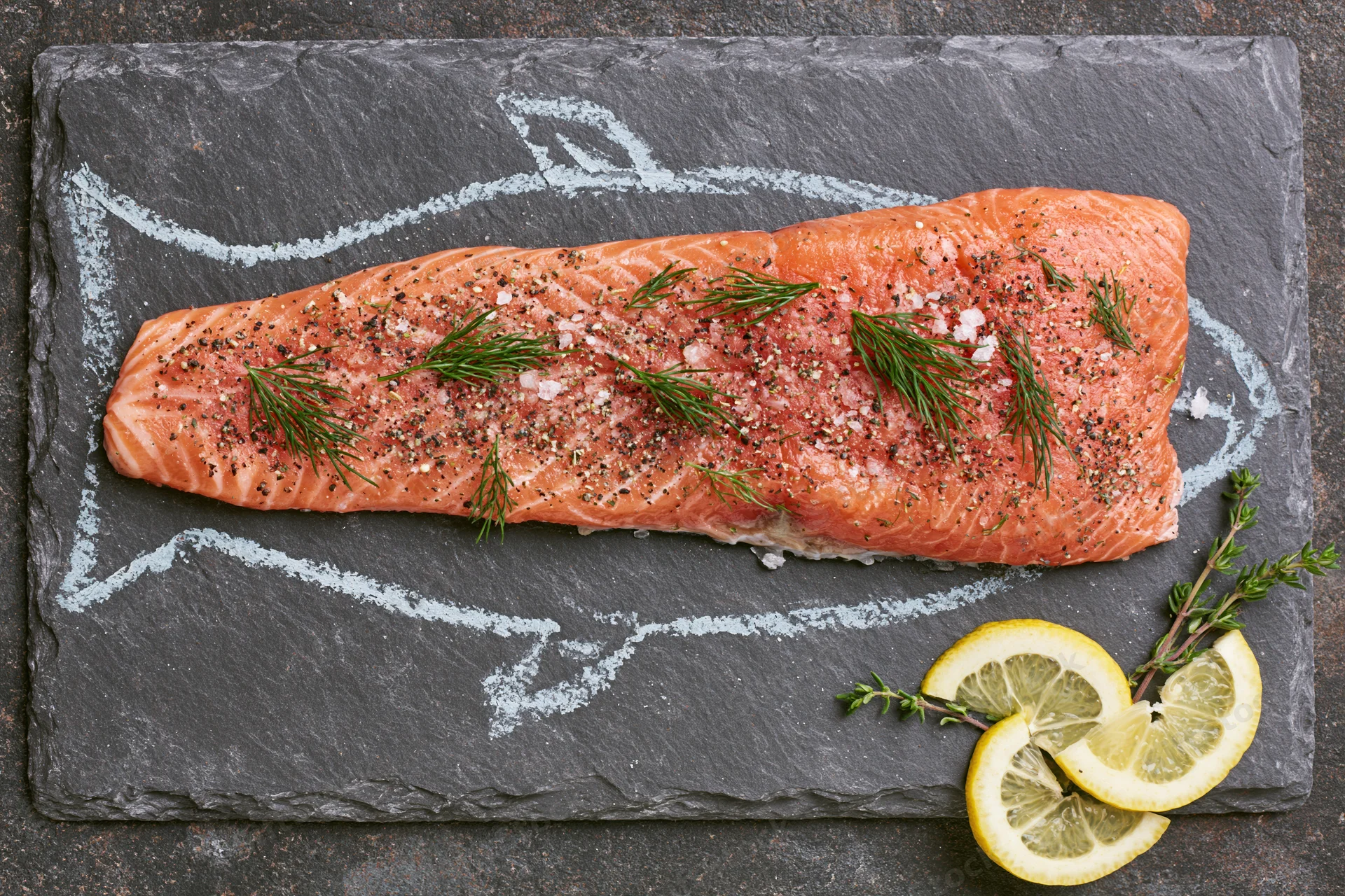 Raw salmon fillet seasoned with dill and pepper on slate plate with lemon garnish