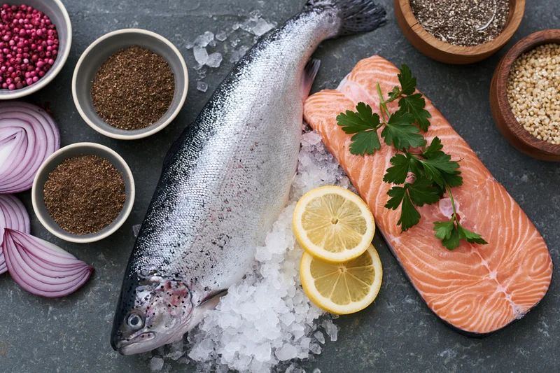 Whole raw salmon on crushed ice beside a fillet with lemon and parsley, surrounded by spices and onions on a gray countertop