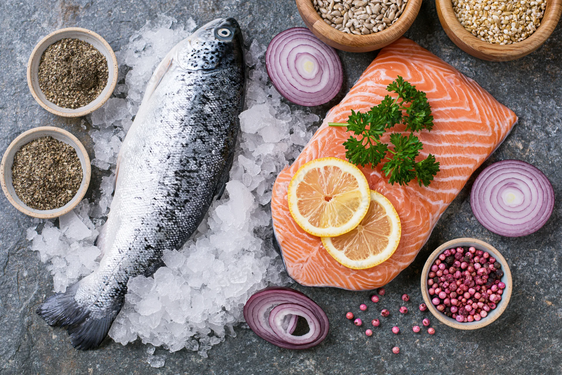 Whole raw fish and salmon fillet on crushed ice with herbs, lemon, onions, and spices arranged on gray surface