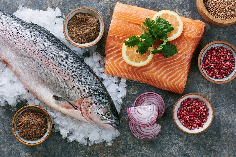 Whole raw salmon on crushed ice beside a fillet garnished with lemon and parsley, surrounded by spices and sliced onion on a dark slate surface