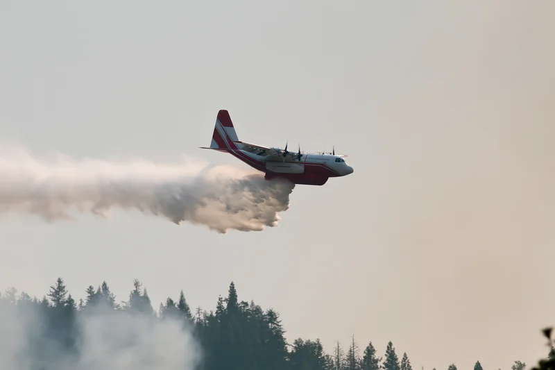 Red and white turboprop airtanker releasing smoke over forest during low flight