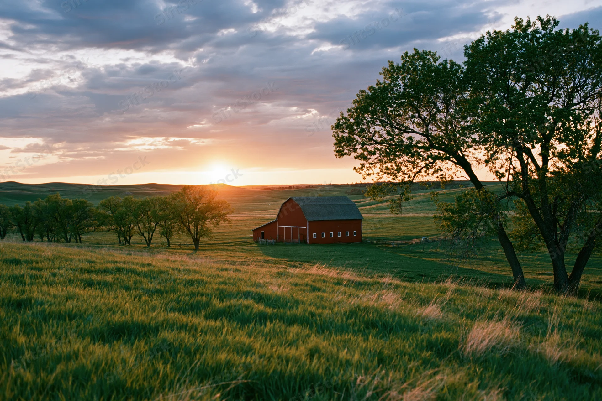 Red barn in a green pasture at sunset with trees and rolling hills under a cloudy sky