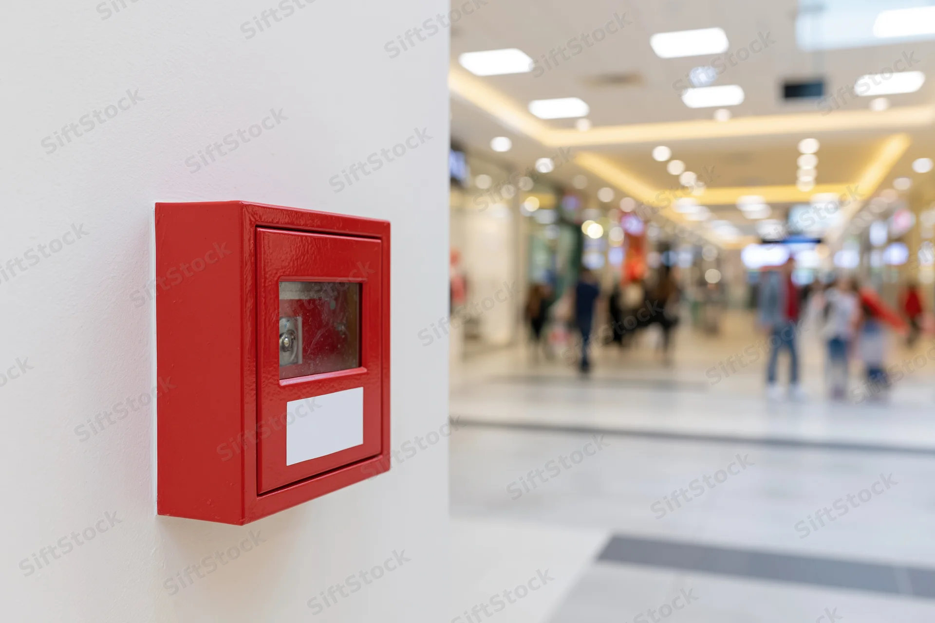 Red fire alarm pull station mounted on white wall in shopping mall corridor