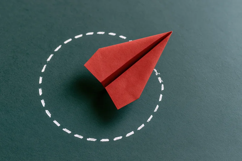 Red origami paper airplane launching upward to the right on dark surface with white dashed circle