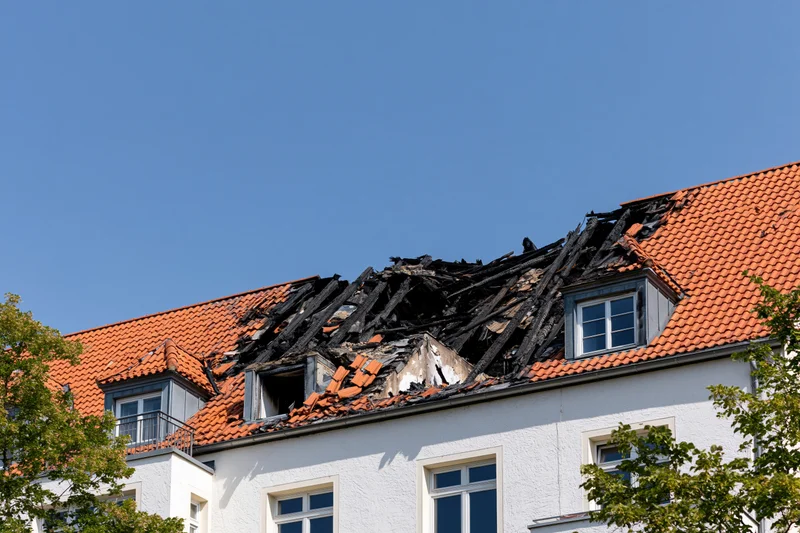 Fire-damaged roof of a residential apartment building with burnt tiles and exposed beams under a clear blue sky