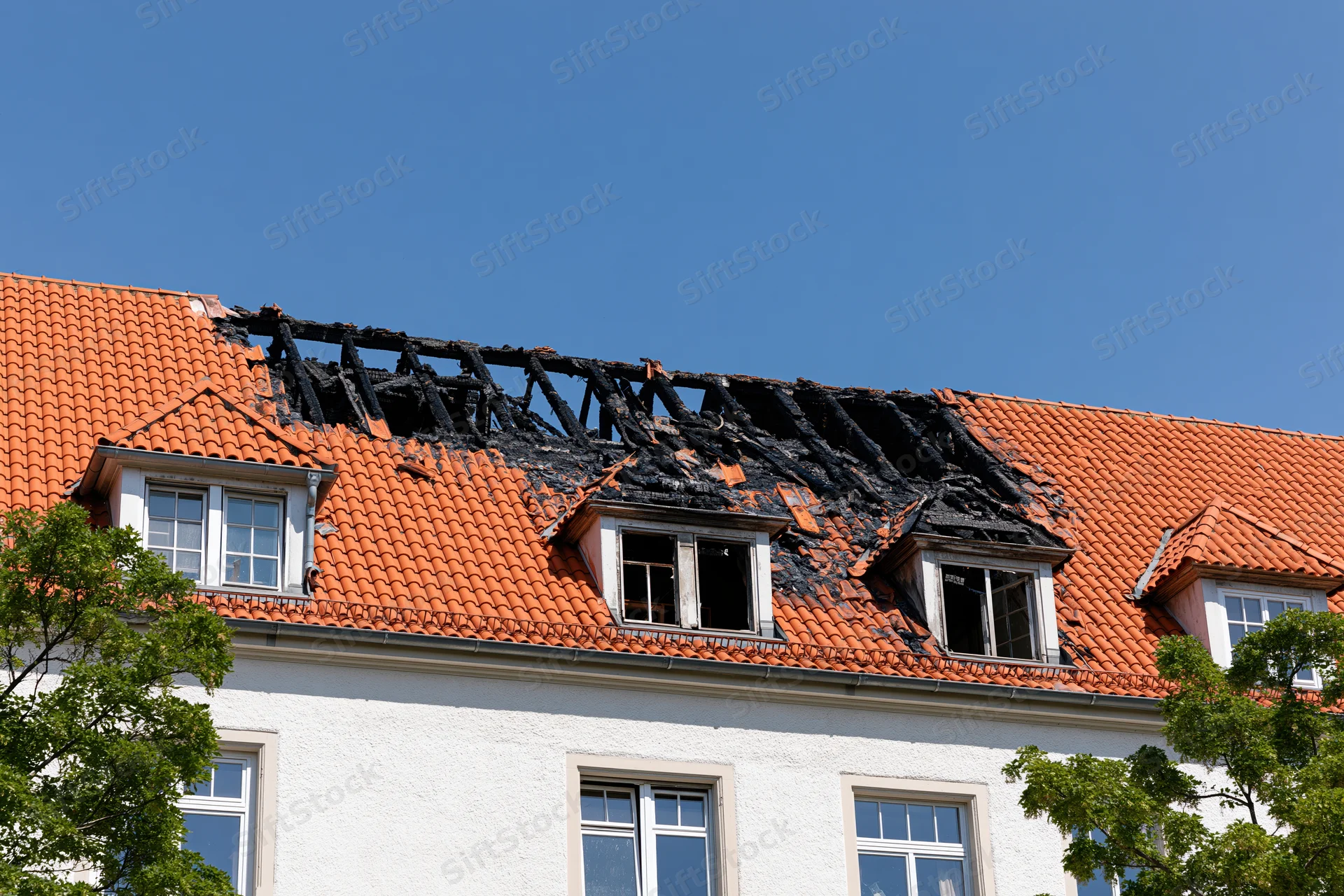 Fire-damaged roof of a residential apartment building with charred beams and broken dormer windows under a clear blue sky