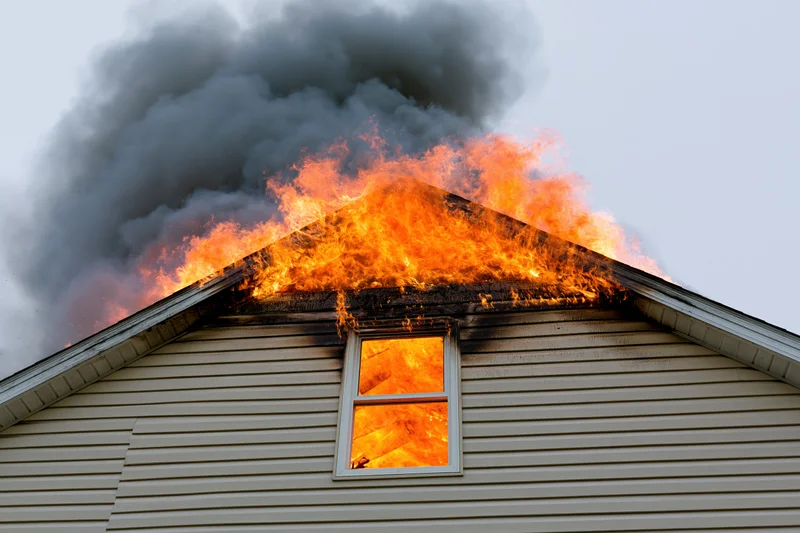 Burning residential house with flames engulfing roof and smoke billowing from attic window