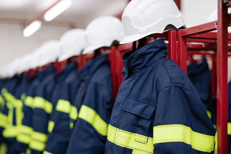 Row of navy blue firefighter turnout gear with white helmets hanging on red metal rack in indoor storage area