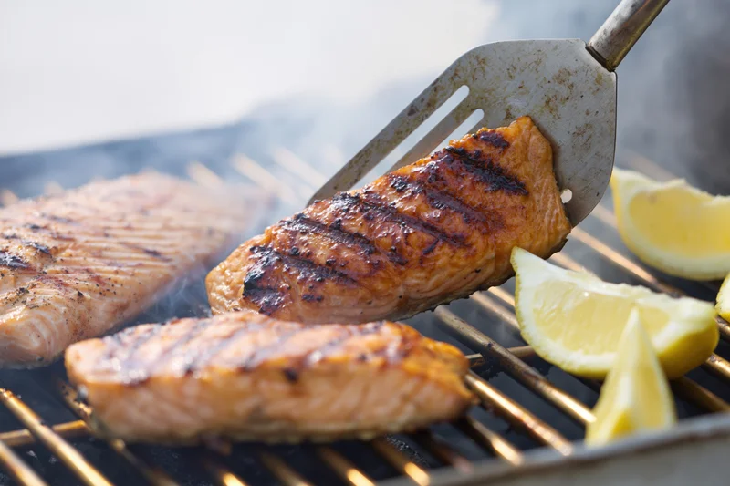 Salmon fillets being flipped with a metal spatula on a hot grill with lemon wedges nearby