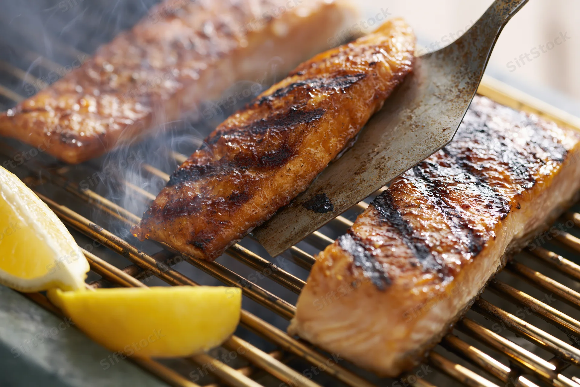 Salmon fillets being flipped with a metal spatula on a hot grill with lemon wedges nearby