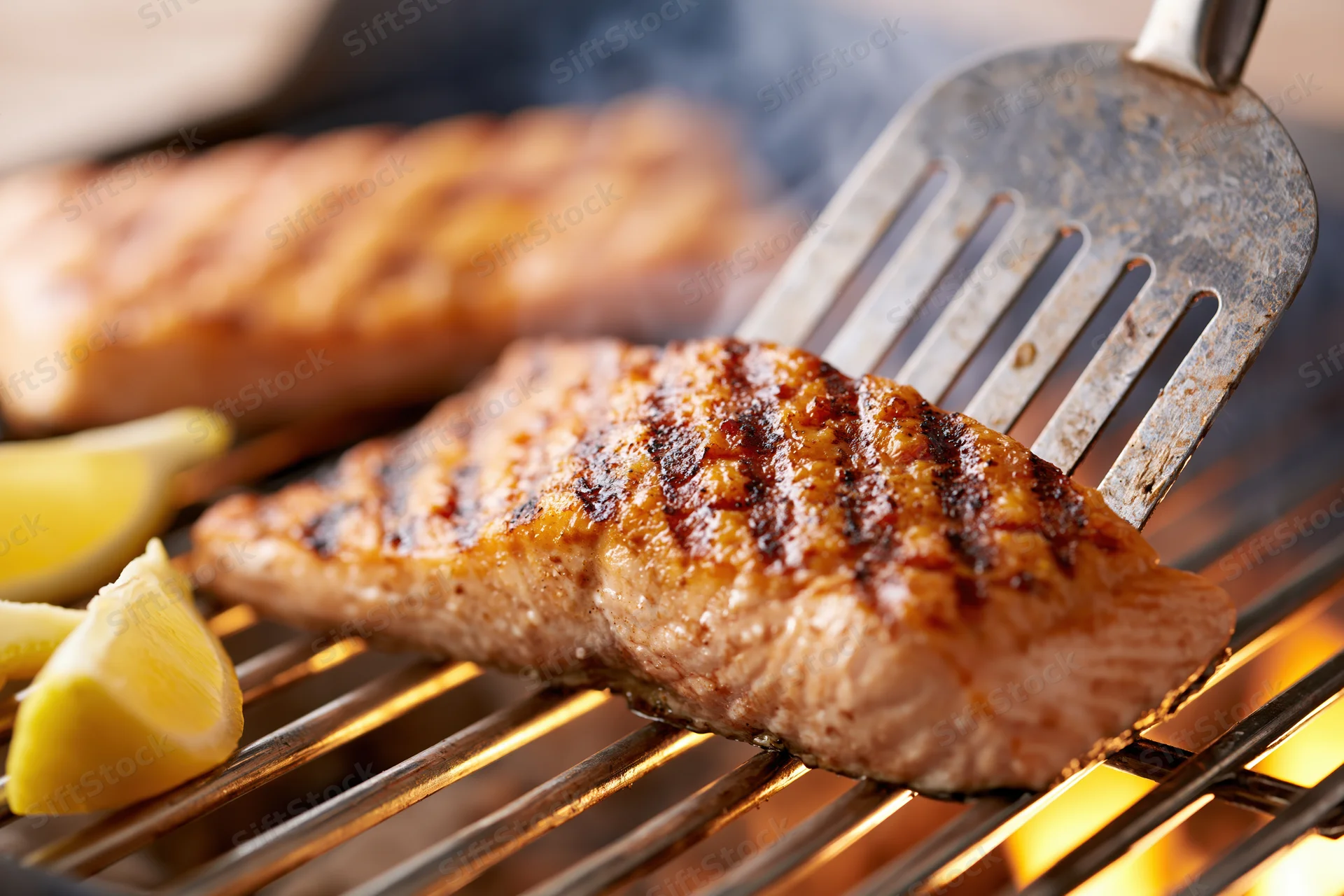 Salmon fillet being flipped with metal spatula on hot grill with lemon wedges nearby