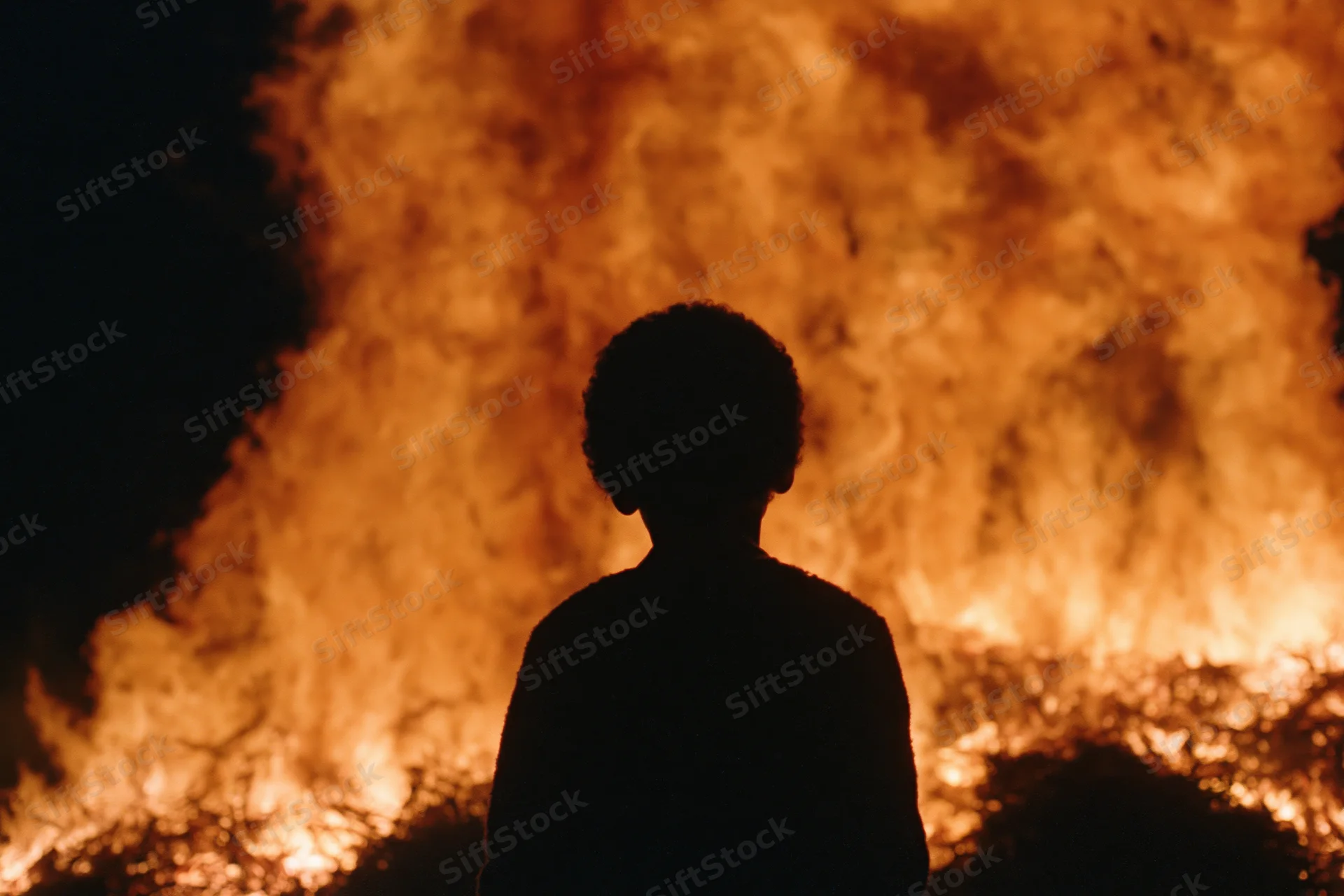 Silhouette of a child standing still facing a massive fire at night