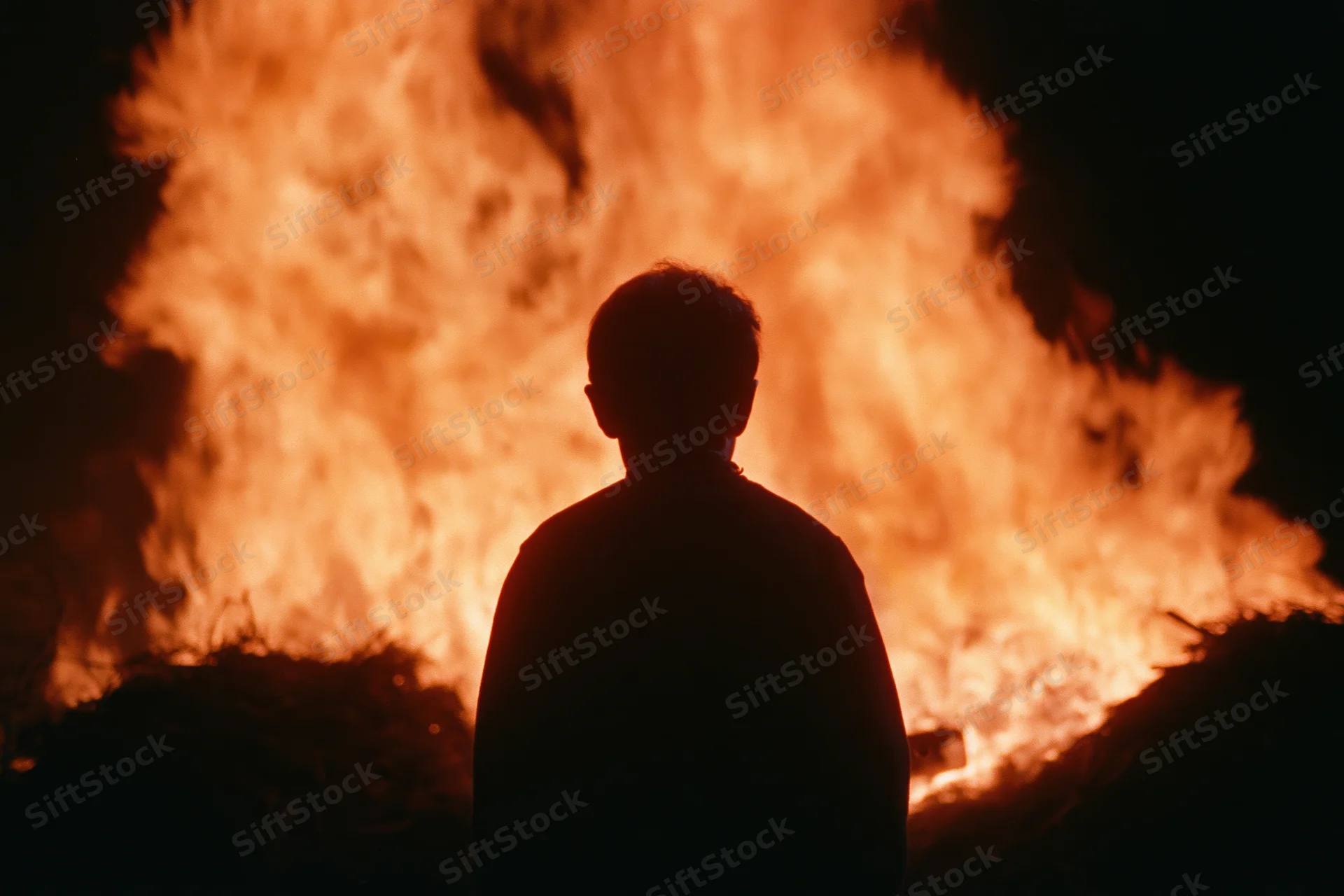 Silhouette of a young child standing still facing a massive fire at night