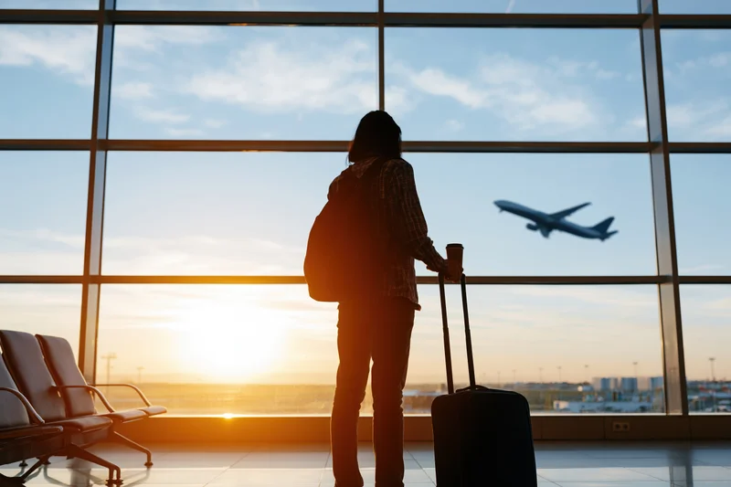 Silhouetted woman with suitcase and backpack standing at airport window watching airplane take off at sunset