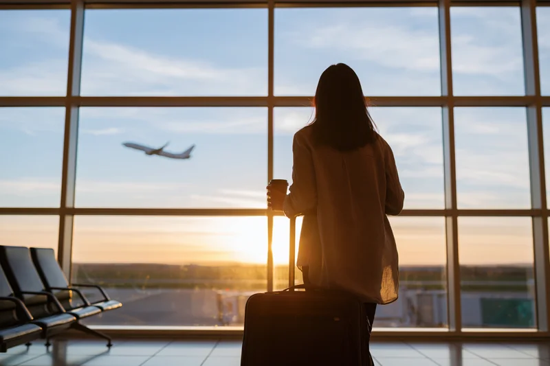 Woman standing alone at airport window holding suitcase and coffee cup watching airplane depart at sunset