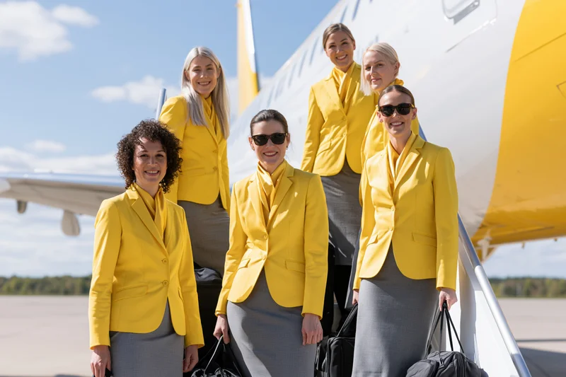 Six flight attendants in yellow blazers and grey skirts standing on tarmac beside airplane smiling at camera