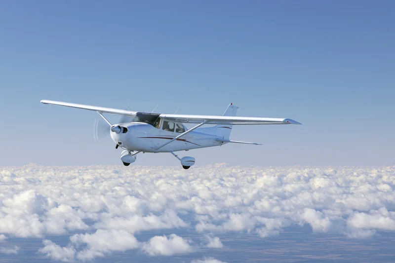 Small white single-engine propeller plane in level flight above cloud layer under clear blue sky