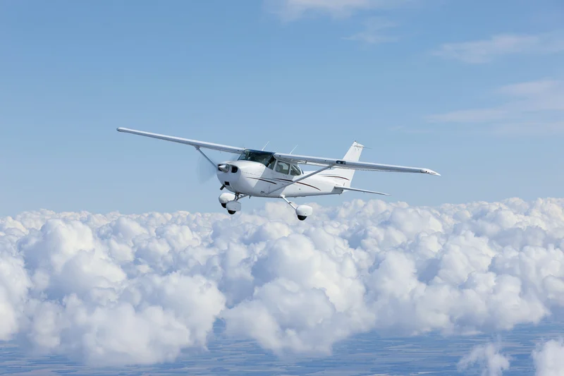 White single-engine propeller airplane in level flight above dense clouds in clear blue sky