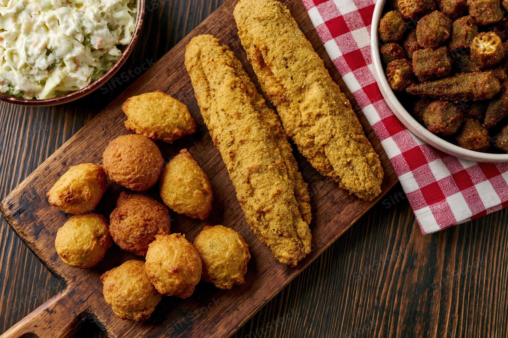 Fried fish and hushpuppies on a wooden serving board with coleslaw and fried okra beside a red checkered napkin