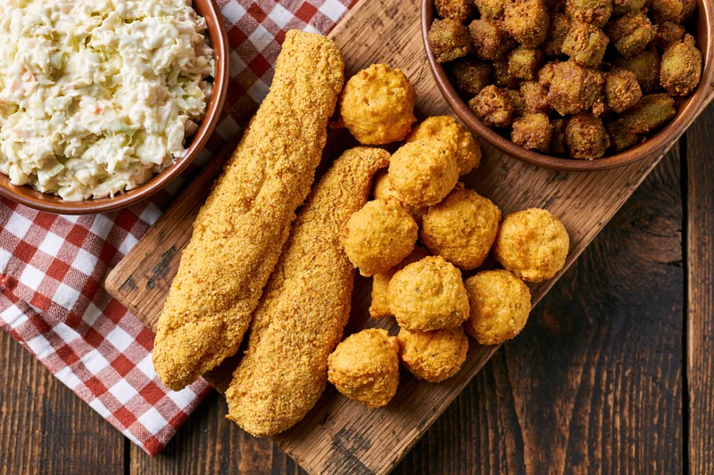 Southern fish fry spread with fried fish sticks, hushpuppies, and coleslaw on a wooden serving board over a checkered cloth