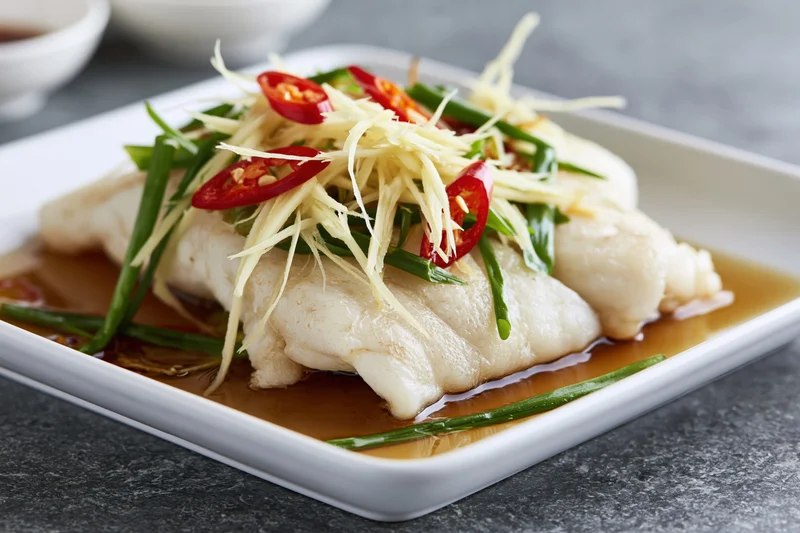Steamed white fish fillet garnished with ginger chili and green onions on a square white ceramic plate