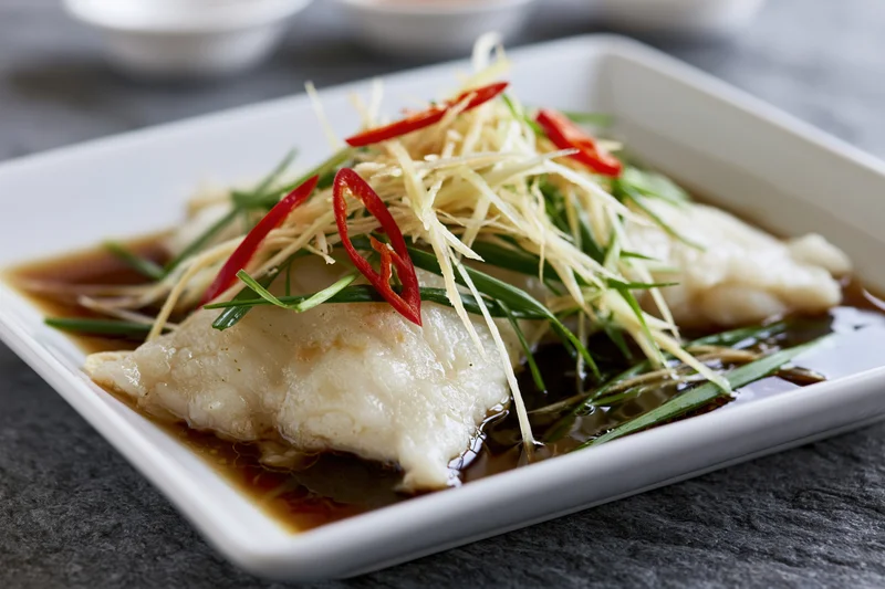 Steamed white fish fillet garnished with ginger, red chili, and green onions on a square white ceramic plate