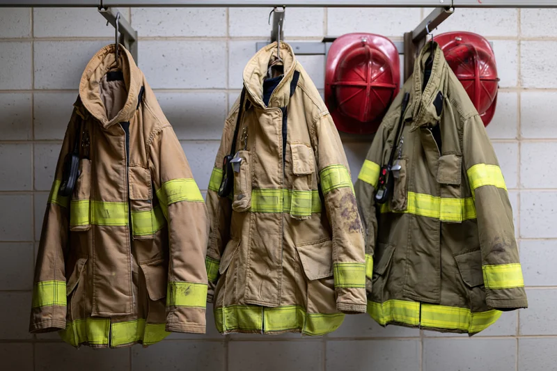Three firefighter turnout coats hanging on a metal rack against a tiled wall