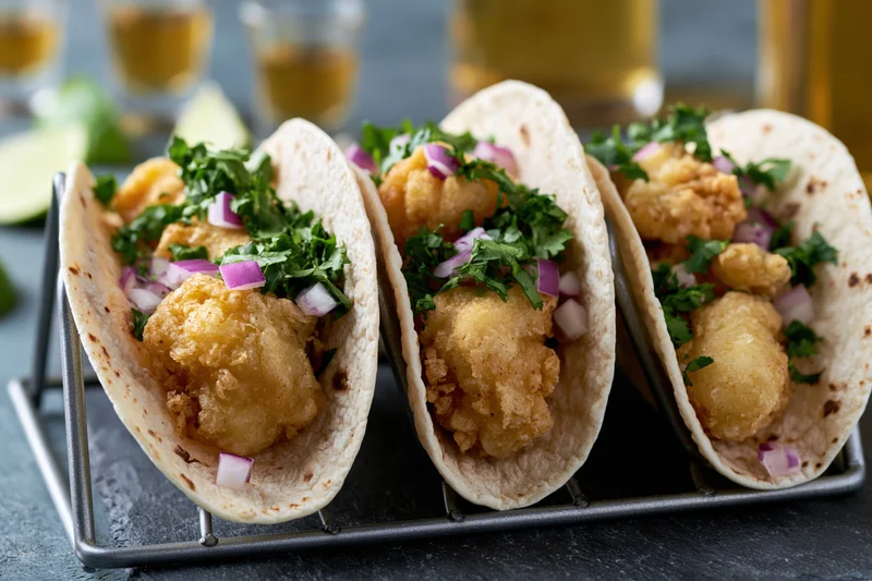 Three fish tacos in flour tortillas held upright in a metal rack with cilantro and red onion garnish