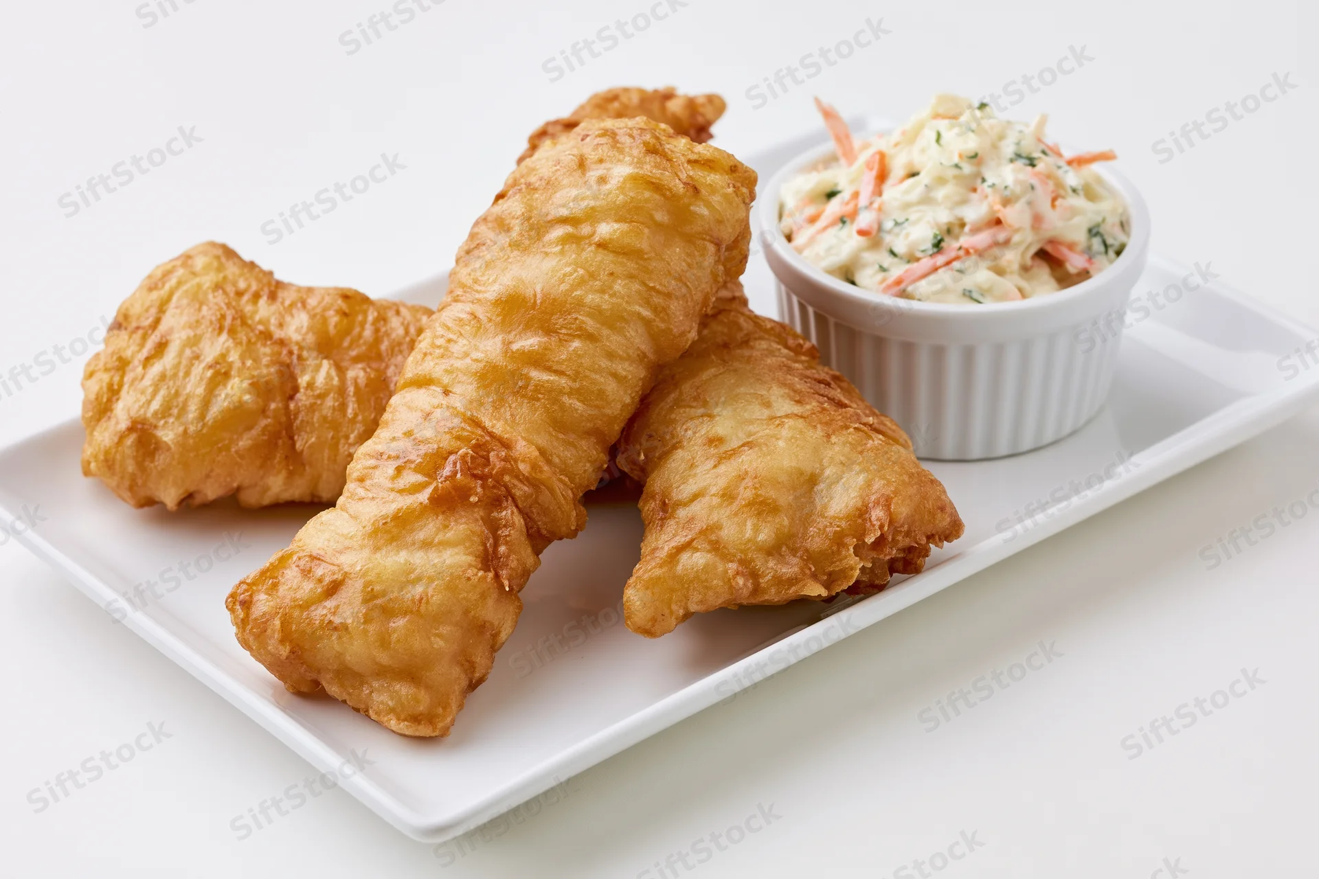 Three golden-brown fried fish fillets with crispy batter on a white plate beside a small bowl of coleslaw