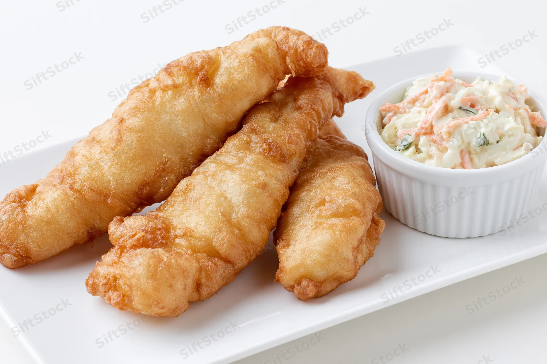 Three golden-brown fried fish fillets with crispy batter on a white plate beside a small bowl of coleslaw