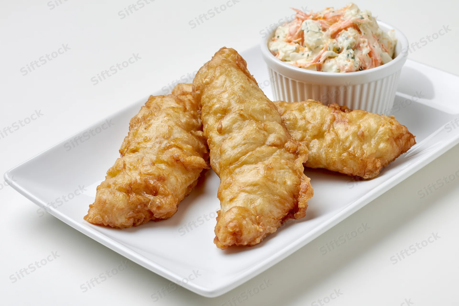 Three golden-brown fried fish fillets with crispy batter on a white plate with a side of coleslaw in a small bowl