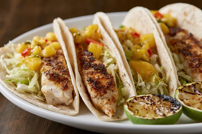 Three grilled fish tacos in flour tortillas filled with shredded cabbage and pineapple salsa on a white plate with grilled lime wedges
