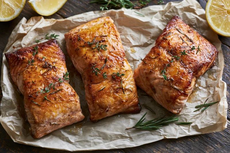 Three pan-seared salmon fillets with rosemary and lemon on parchment paper on a wooden table