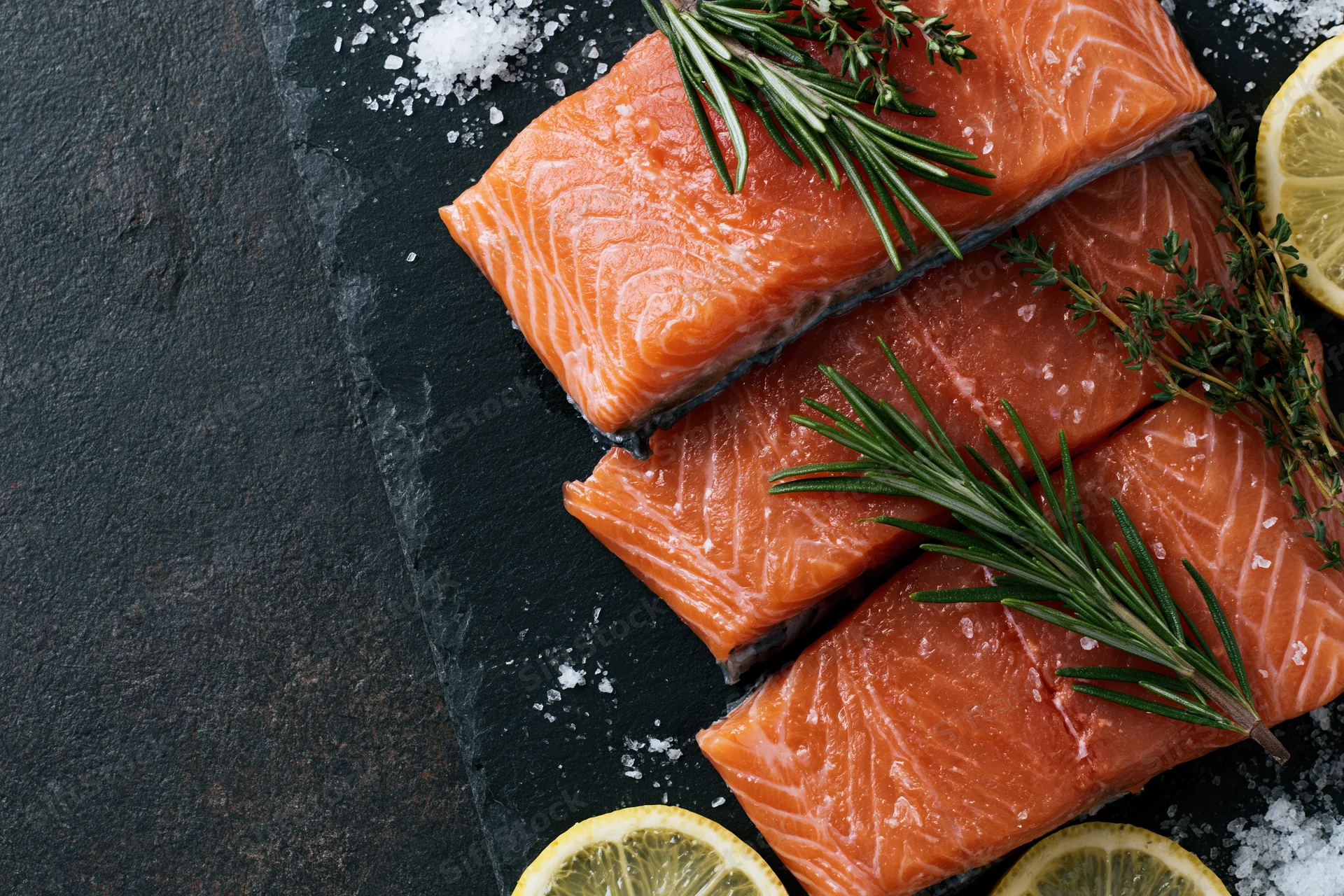 Three raw salmon fillets arranged on dark slate with herbs, lemon slices and salt