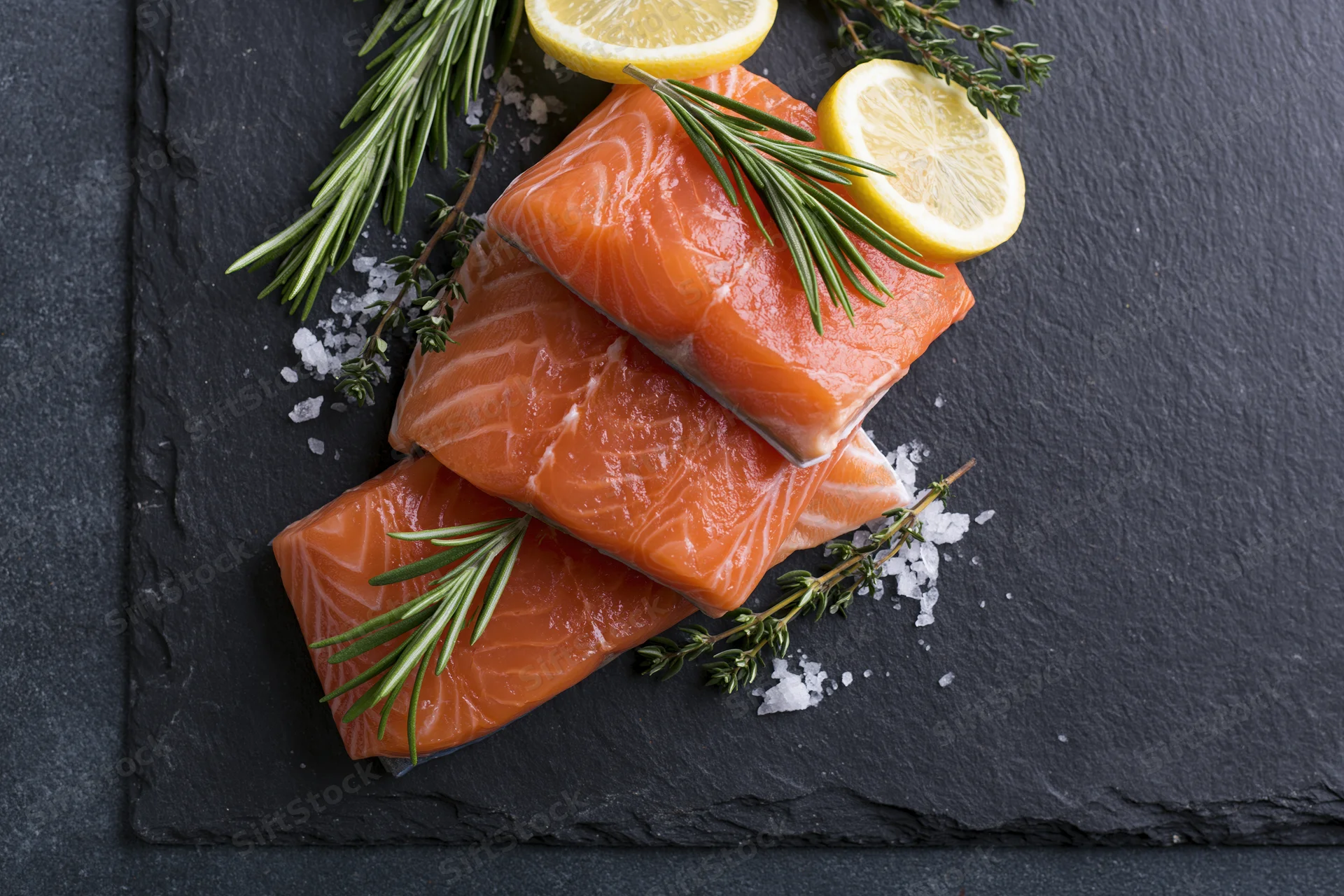 Three raw salmon fillets stacked on a dark slate board with lemon slices, rosemary, thyme, and coarse salt