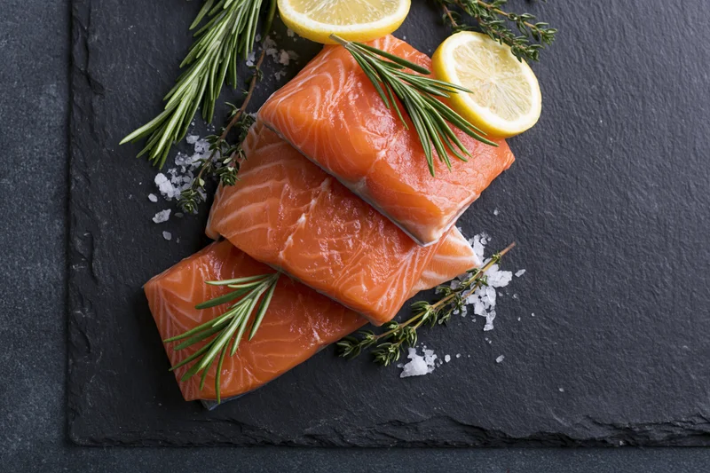 Three raw salmon fillets stacked on a dark slate board with lemon slices, rosemary, thyme, and coarse salt