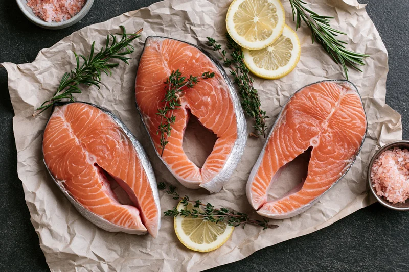 Three raw salmon steaks arranged on crinkled parchment paper with herbs, lemon slices, and salt bowls