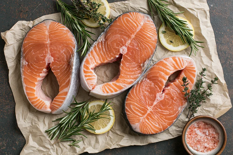 Three raw salmon steaks arranged on crumpled parchment paper with herbs, lemon slices, and salt
