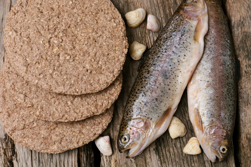 Two raw whole trout beside stacked whole wheat flatbreads on a wooden surface