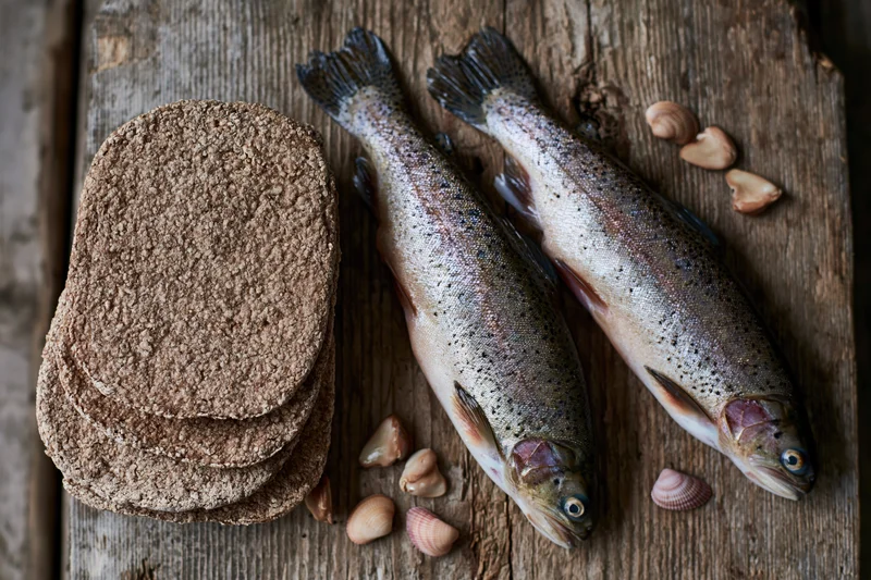 Two raw whole trout beside a stack of whole wheat flatbreads on a wooden table