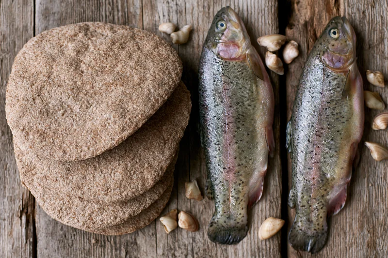 Two raw whole trout beside a stack of whole wheat flatbreads on a wooden surface