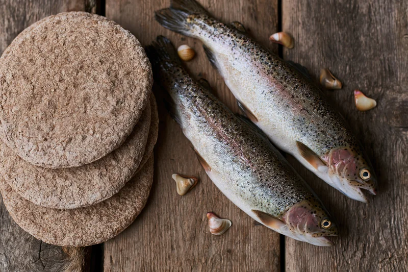 Two raw whole trout beside a stack of whole wheat flatbreads on a wooden table