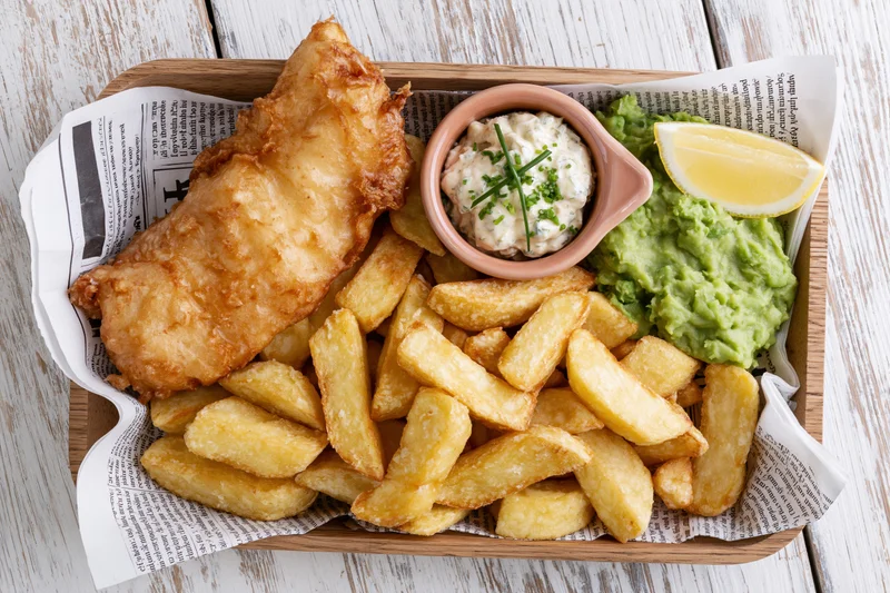 Golden fried fish and thick-cut chips served on newspaper-lined tray with tartar sauce, guacamole, and lemon wedge