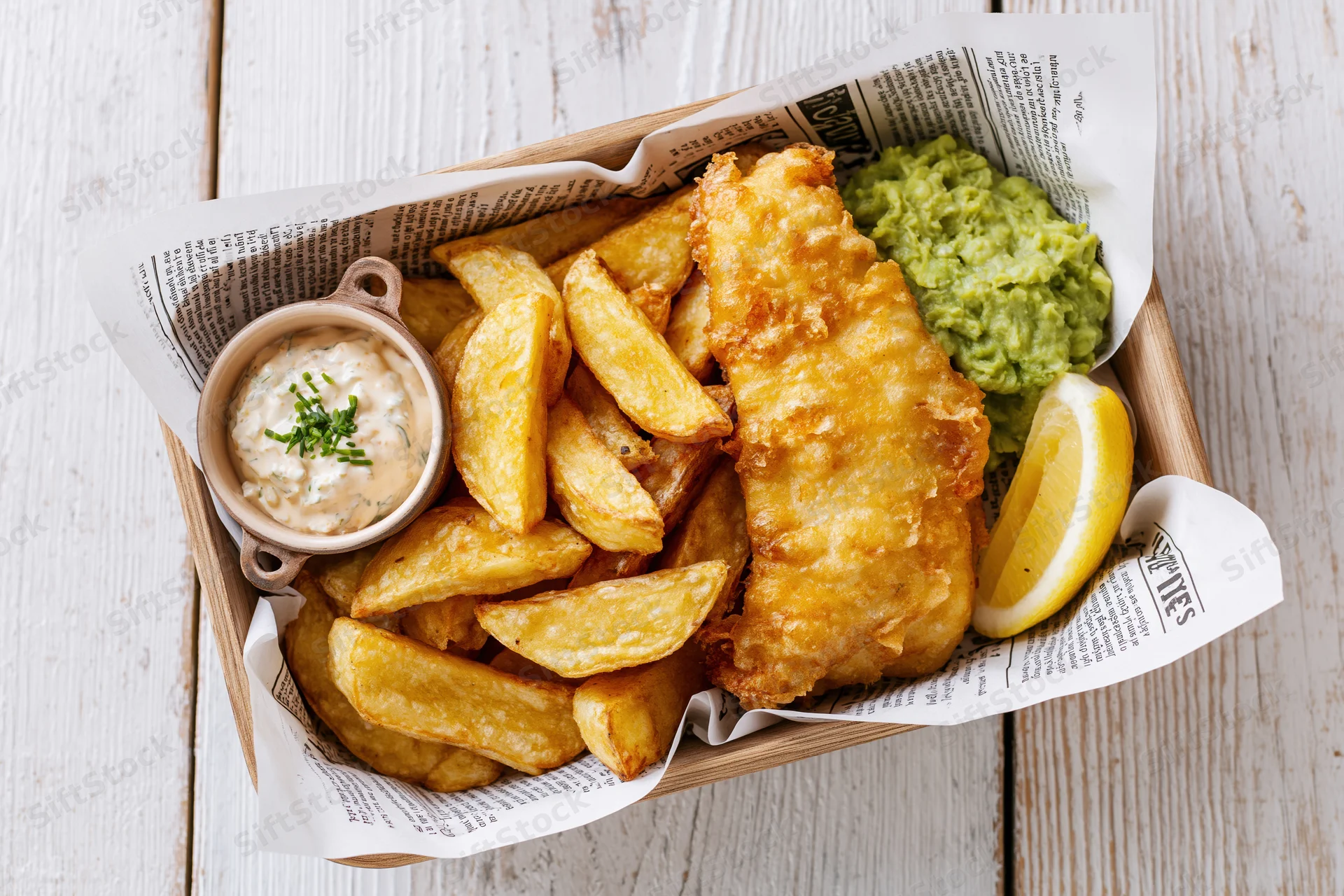 Fish and chips served in a newspaper-lined wooden basket with tartar sauce, lemon, and mushy peas on a white wooden table