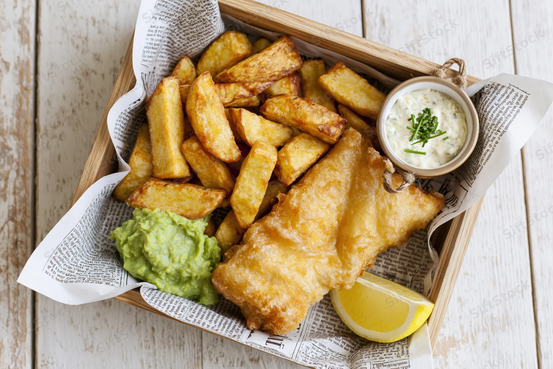 Golden fried fish and thick-cut chips served in a newspaper-lined wooden tray with tartar sauce, lemon wedge, and mushy peas