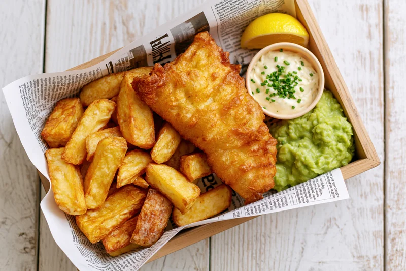 Golden fried fish and thick-cut chips served in a newspaper-lined wooden tray with mushy peas, tartar sauce and lemon wedge