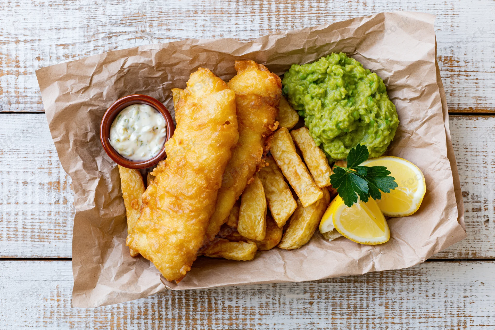Fish and chips served on crinkled brown paper with tartar sauce, lemon, parsley, and guacamole on a rustic white wooden table