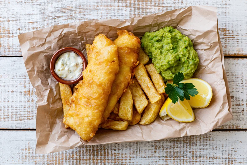 Fish and chips served on crinkled brown paper with tartar sauce, lemon, parsley, and guacamole on a rustic white wooden table