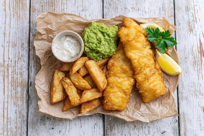 Two pieces of battered fish with thick-cut chips, tartar sauce, guacamole and lemon wedges on crinkled brown paper over a white wooden table